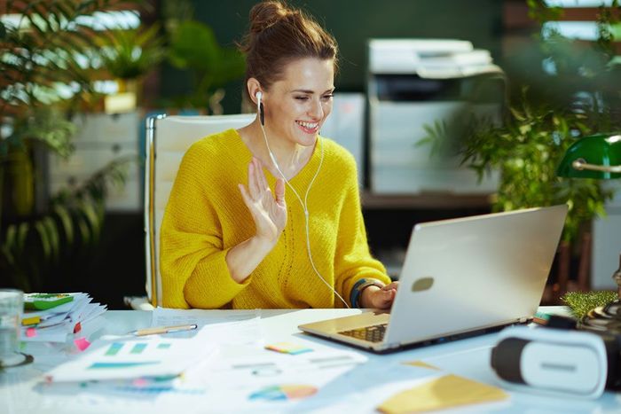 Woman with laptop waves, wearing yellow sweater and earphones, indoors, smiling.