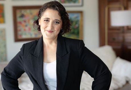 Woman in a black blazer and white top, smiling, hands on hips, in a home setting.