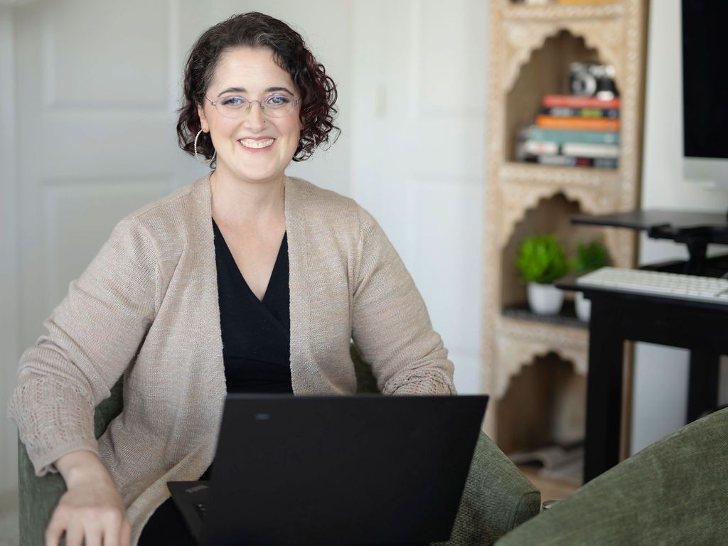 Woman smiles while sitting with a laptop. She wears glasses and a tan cardigan in a home office.