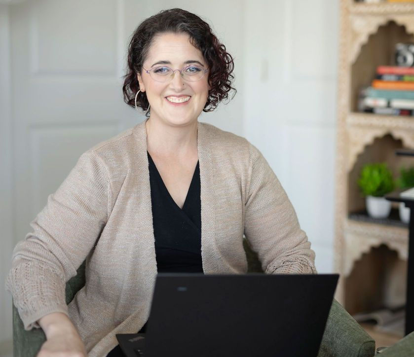 Woman seated with laptop, smiling. Wearing glasses, cardigan, near a bookshelf.