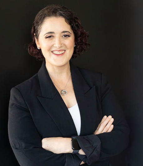 Woman with short curly hair, smiling, wearing a black blazer and white top, arms crossed, black background.