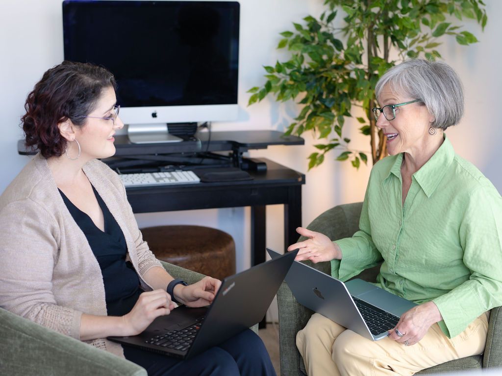 Two women seated, each with a laptop, conversing in an office setting. One points while the other listens.