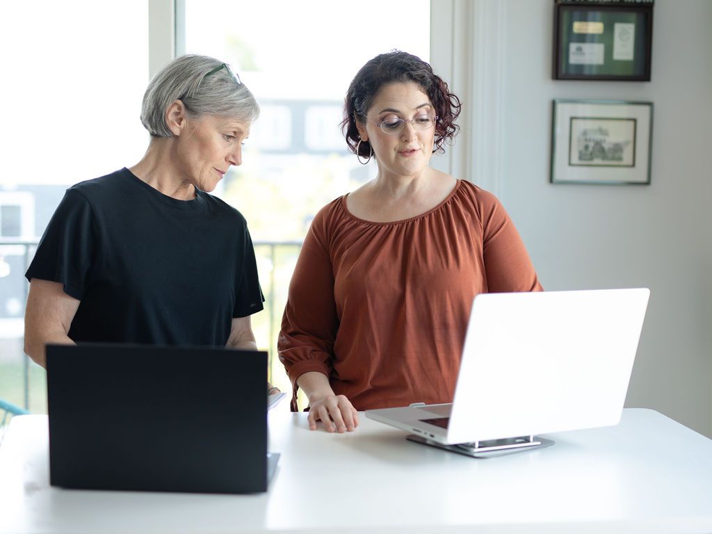 Two women looking at laptop screens at a white table in a bright room.