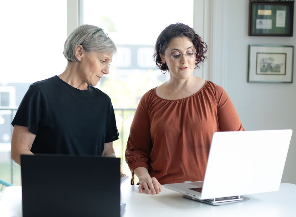 Two women looking at laptop screens. One in black shirt, the other in orange, in a bright room.