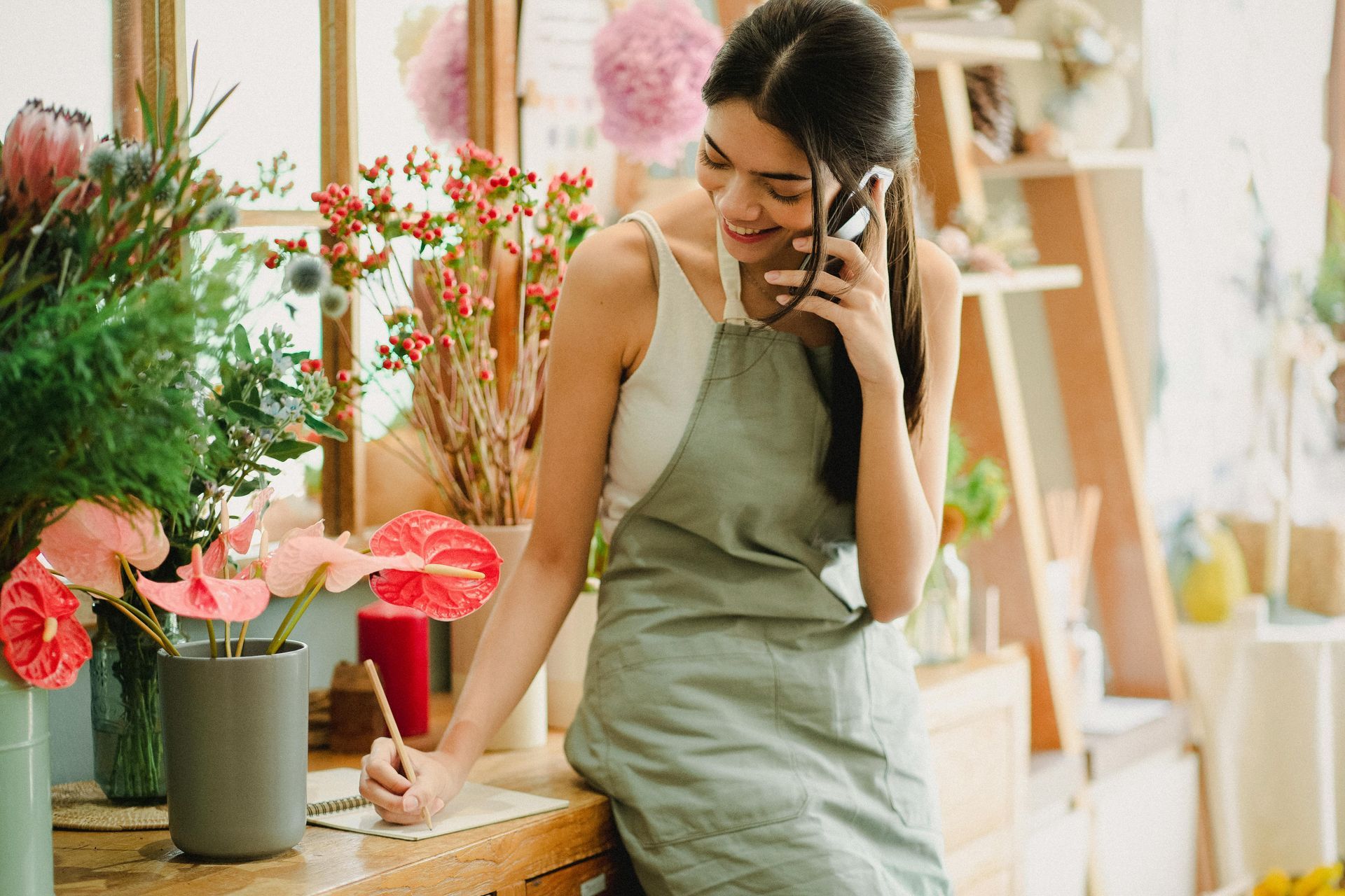 Woman in an apron, smiling while on a phone call, writing on a notepad in a flower shop.