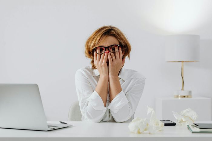 Woman with hands over face, wearing glasses, at a desk with a laptop and crumpled tissues, appearing stressed.