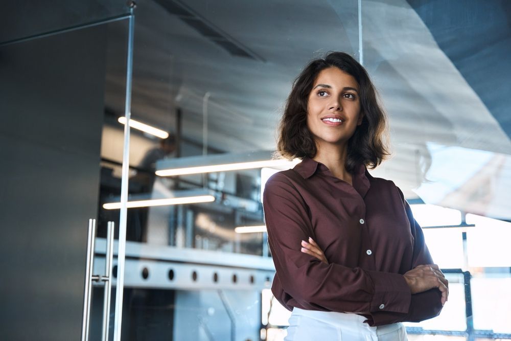 Woman in burgundy shirt, arms crossed, smiles confidently near glass office doors.
