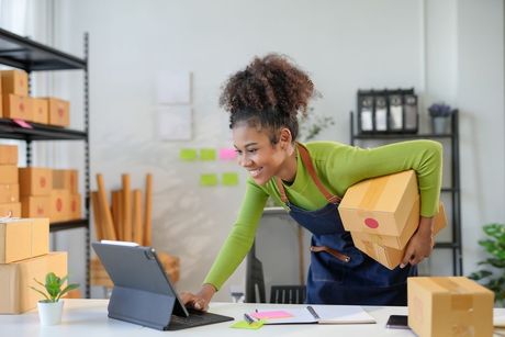 Woman in apron smiling at a tablet, holding boxes. Small business, packing orders.