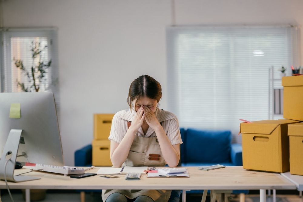 Woman at desk, hands on face, looking stressed. Surrounded by boxes and office supplies.