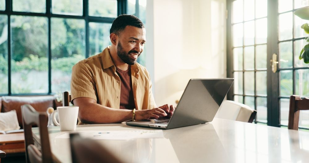 Man smiles while typing on a laptop at a table near a window; coffee cup present.