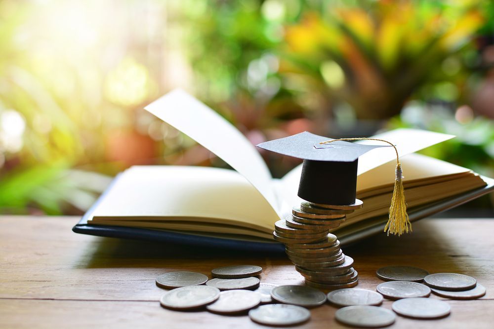 Graduation cap balanced on a stack of coins, atop an open book, scattered coins, outdoors.