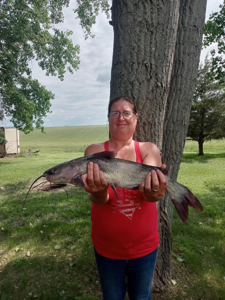 A woman in a red tank top is holding a catfish in front of a tree.