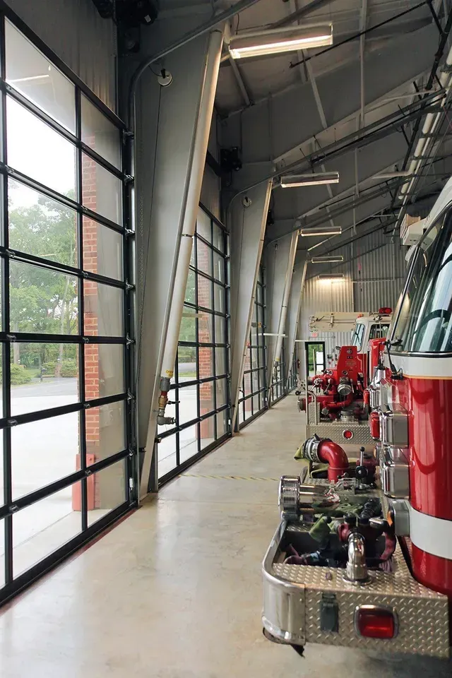 Fire trucks parked in a modern fire station bay with glass doors and a concrete floor.
