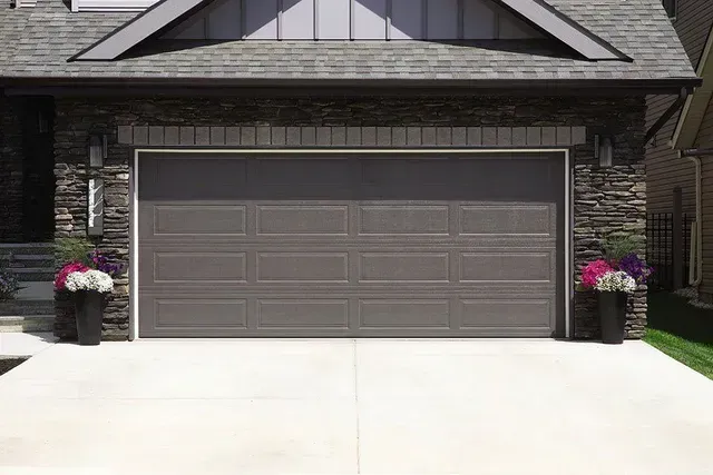 Brown garage door in front of a house, flanked by planters with purple and white flowers.
