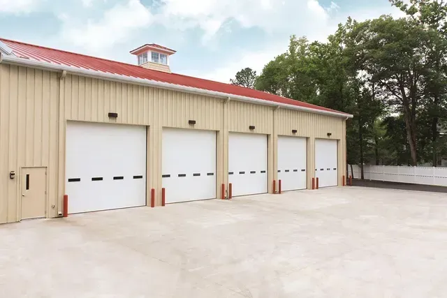 Beige metal building with four white garage doors, red roof, and a small tower.