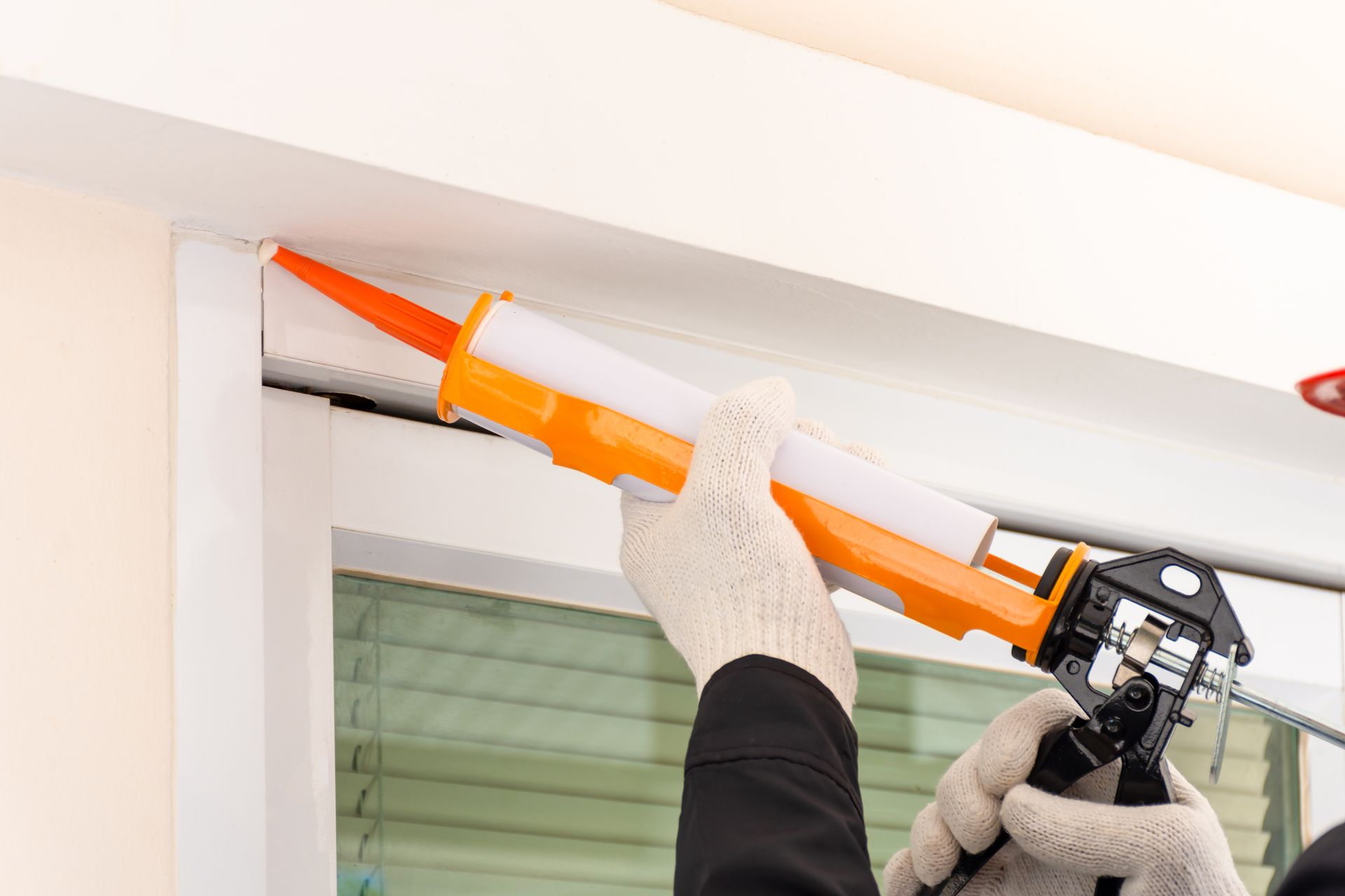 Person wearing gloves caulking a white window frame with an orange and white caulk gun.