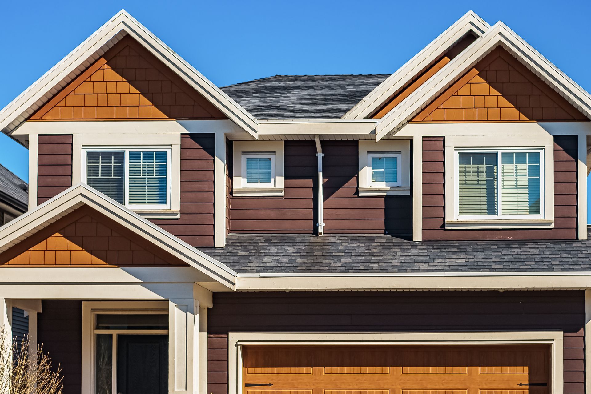 Two-story house with brown siding, orange accents, and a gray shingled roof, against a blue sky.