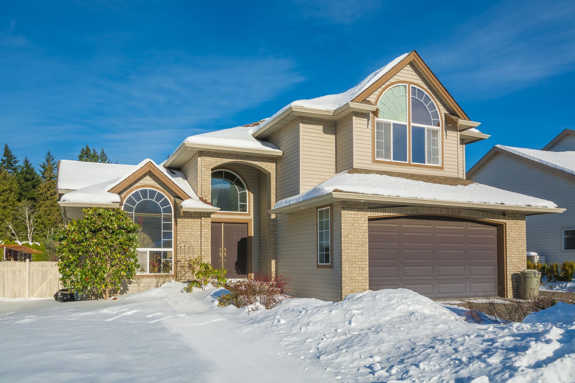Two-story house with snow-covered roof and yard; brown garage door, light brick exterior, and arched windows against blue sky.