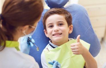 A young boy is sitting in a dental chair giving a thumbs up.