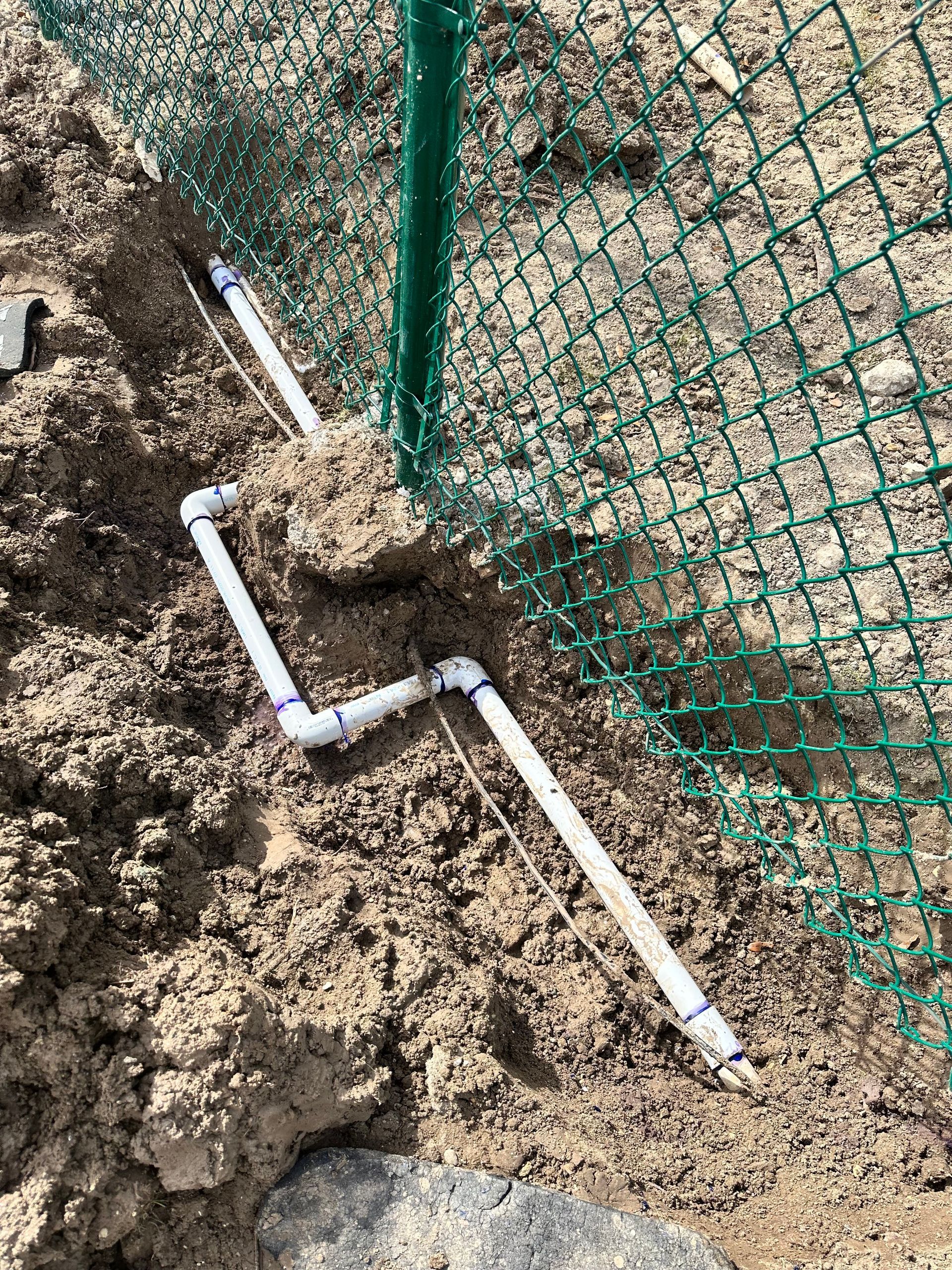 A white pipe is laying in the dirt next to a green fence.