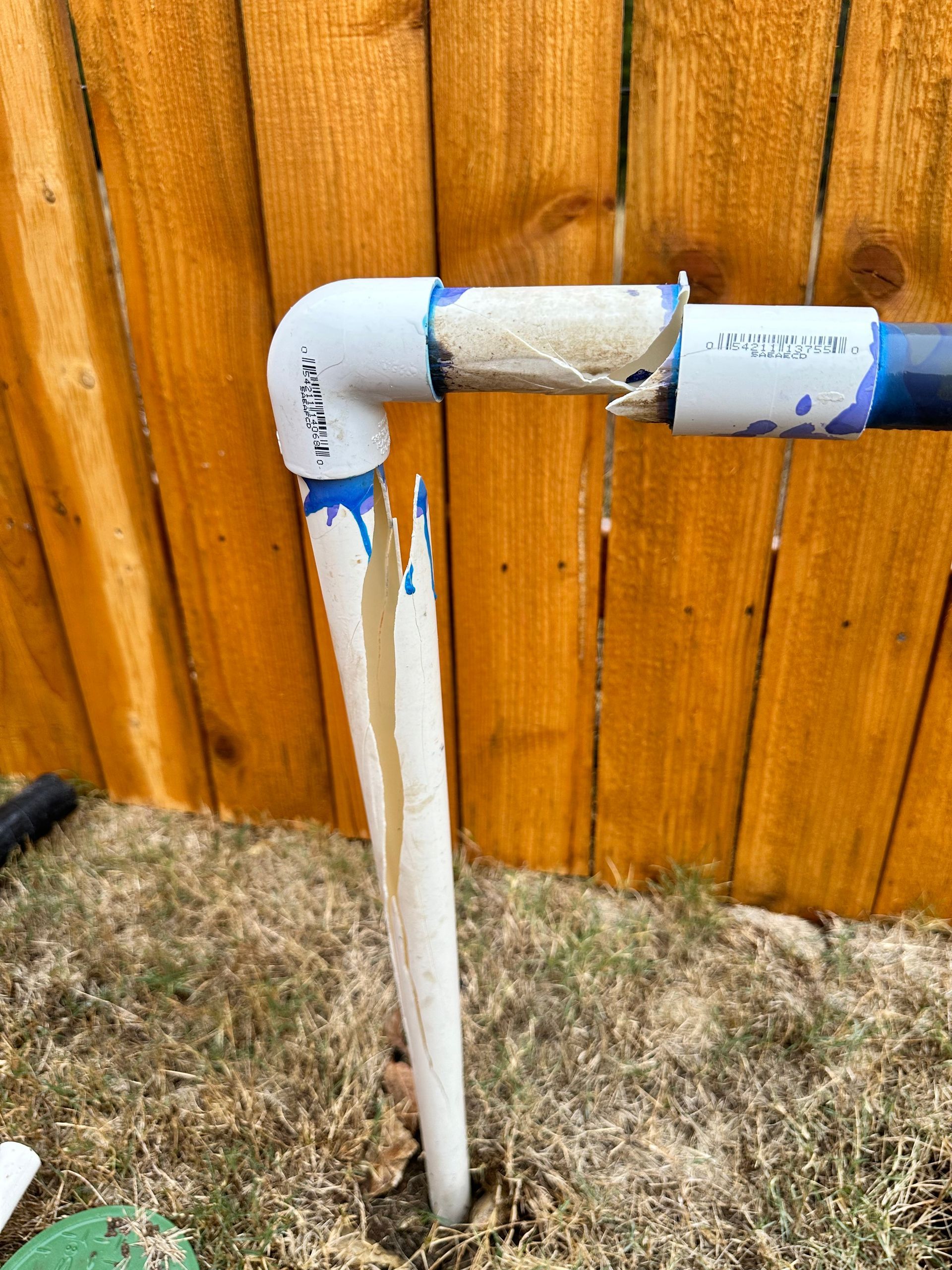 A broken pipe is sitting in the grass in front of a wooden fence.