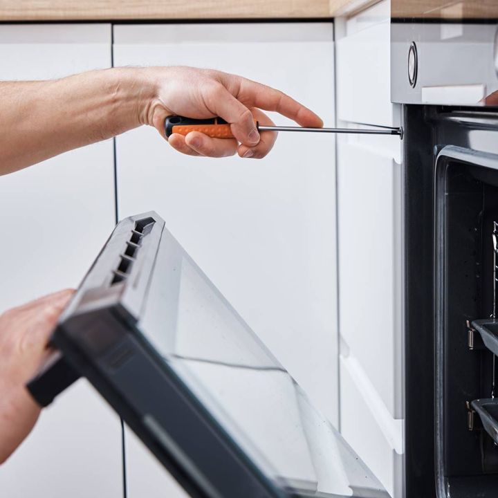 Person using a screwdriver to work on an oven door in a white kitchen.
