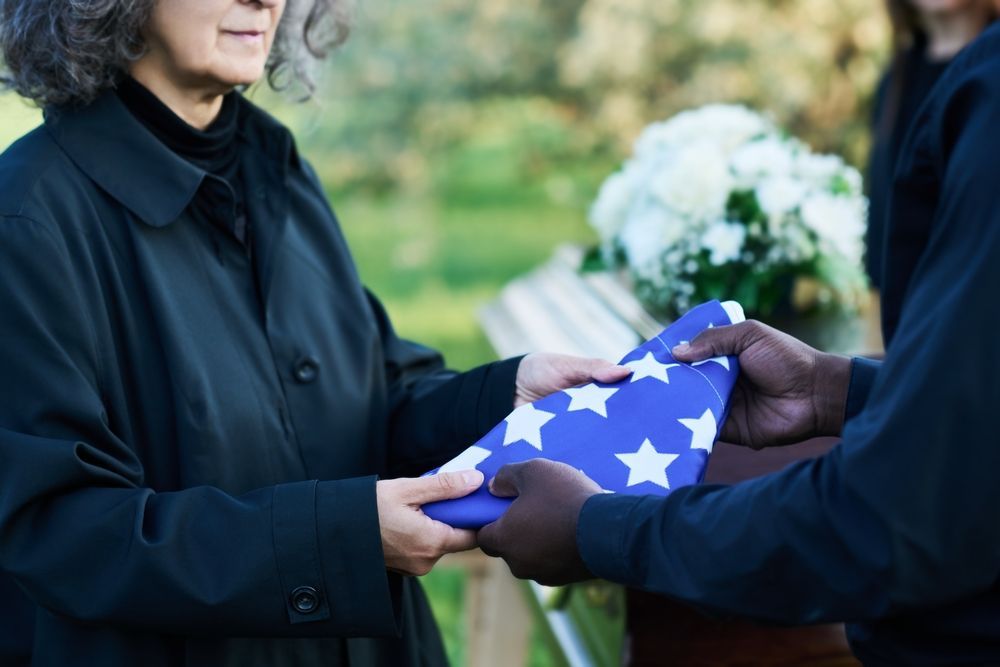 A person receives a folded American flag at a funeral. Black clothing, flowers, and outdoors setting.