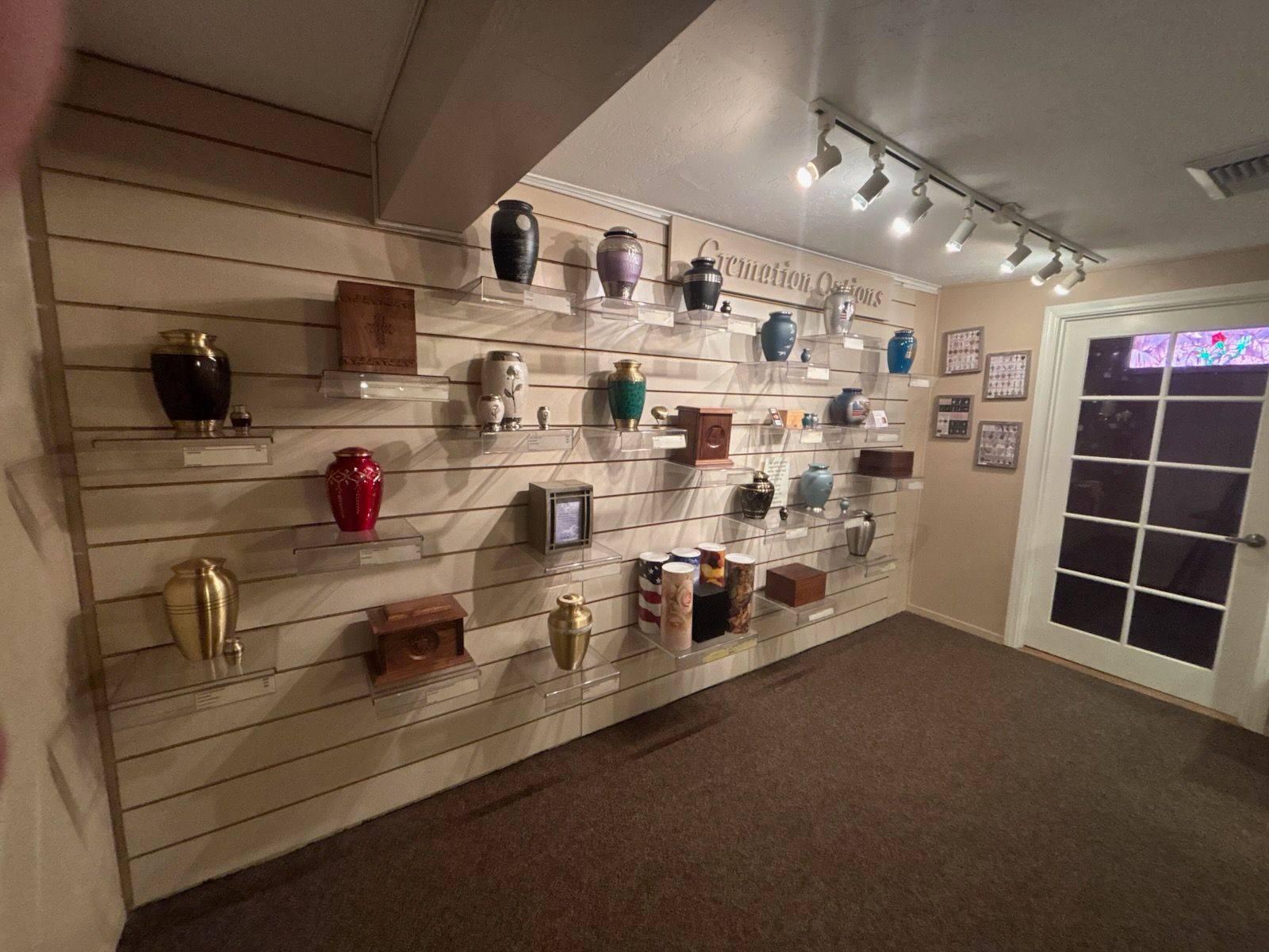 Wall display of cremation urns in various colors and shapes, arranged on shelves in a funeral home showroom.