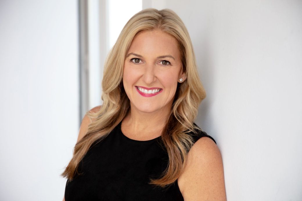 A woman in a black dress is smiling and leaning against a wall.