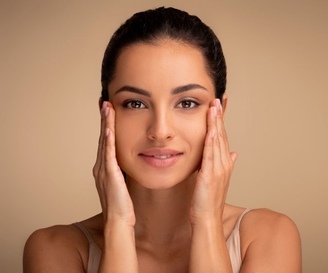 A Woman is Touching Her Face With Her Hands and Smiling — Coastal Skin & Laser in Tewantin, QLD