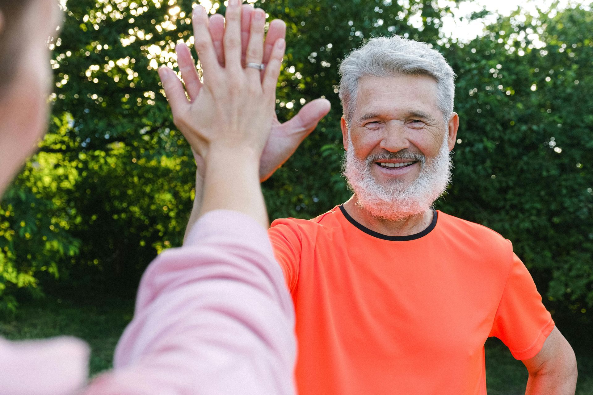 An individual with a white beard in an orange shirt high-fives another person outdoors against a backdrop of green trees.