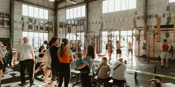 People watching a workout in a gym with high ceilings and large windows. Several people are exercising.