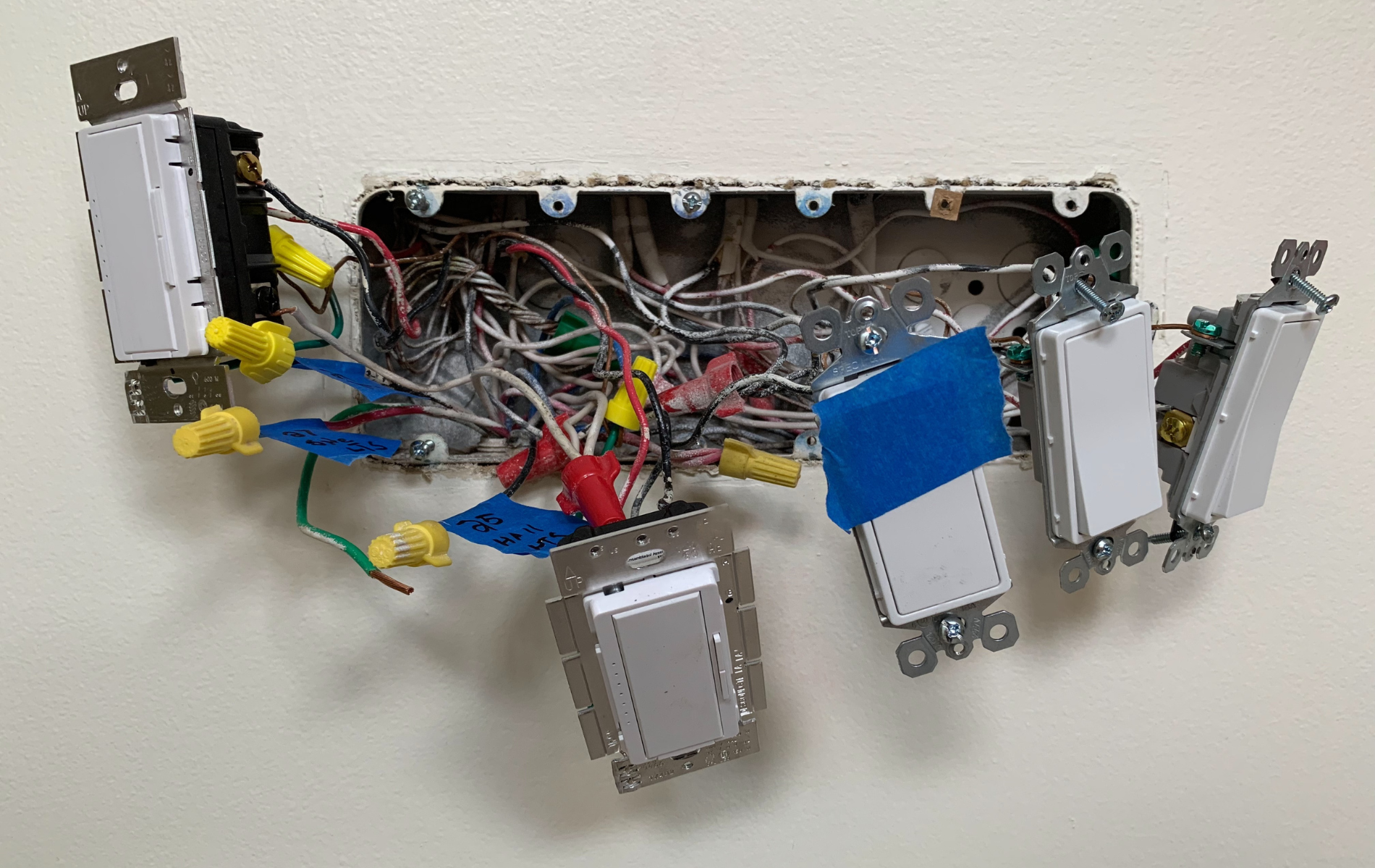 Open electrical box on a wall with several light switches and many wires, various colors, connected with wire nuts.