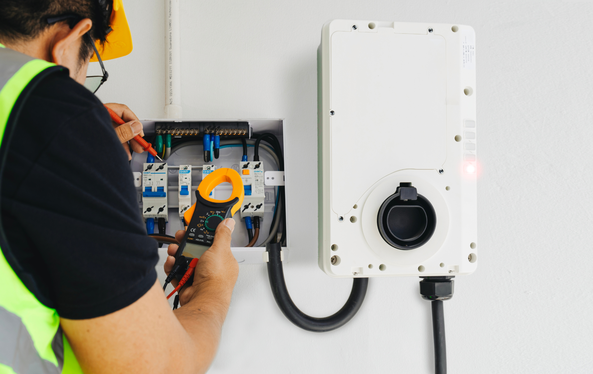 Electrician in safety vest using a multimeter on electrical box next to an EV charger on a white wall.