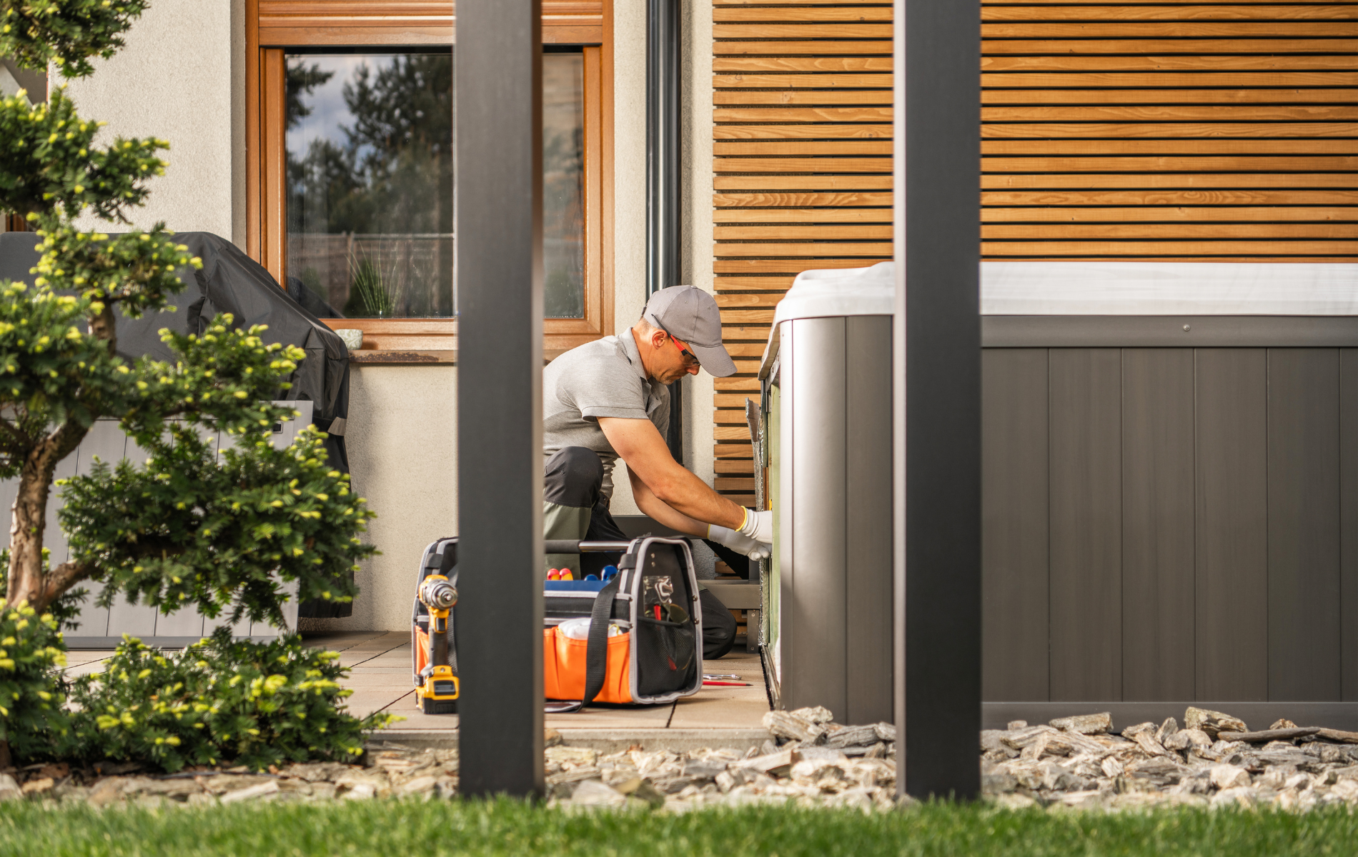 A technician repairs a hot tub outdoors, next to a house with wooden siding.