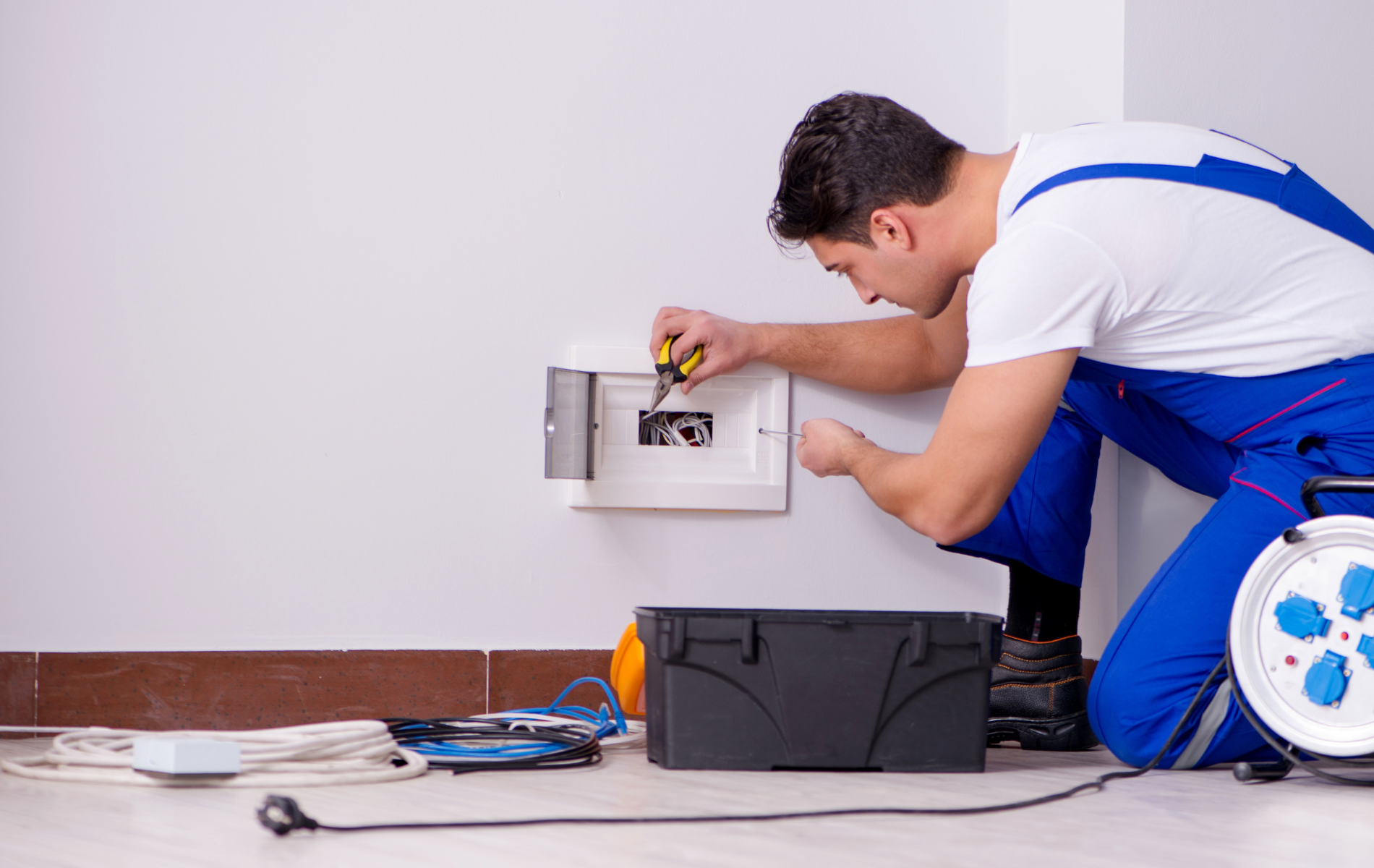 Electrician in blue coveralls installing electrical box, working on wall with tools on floor.