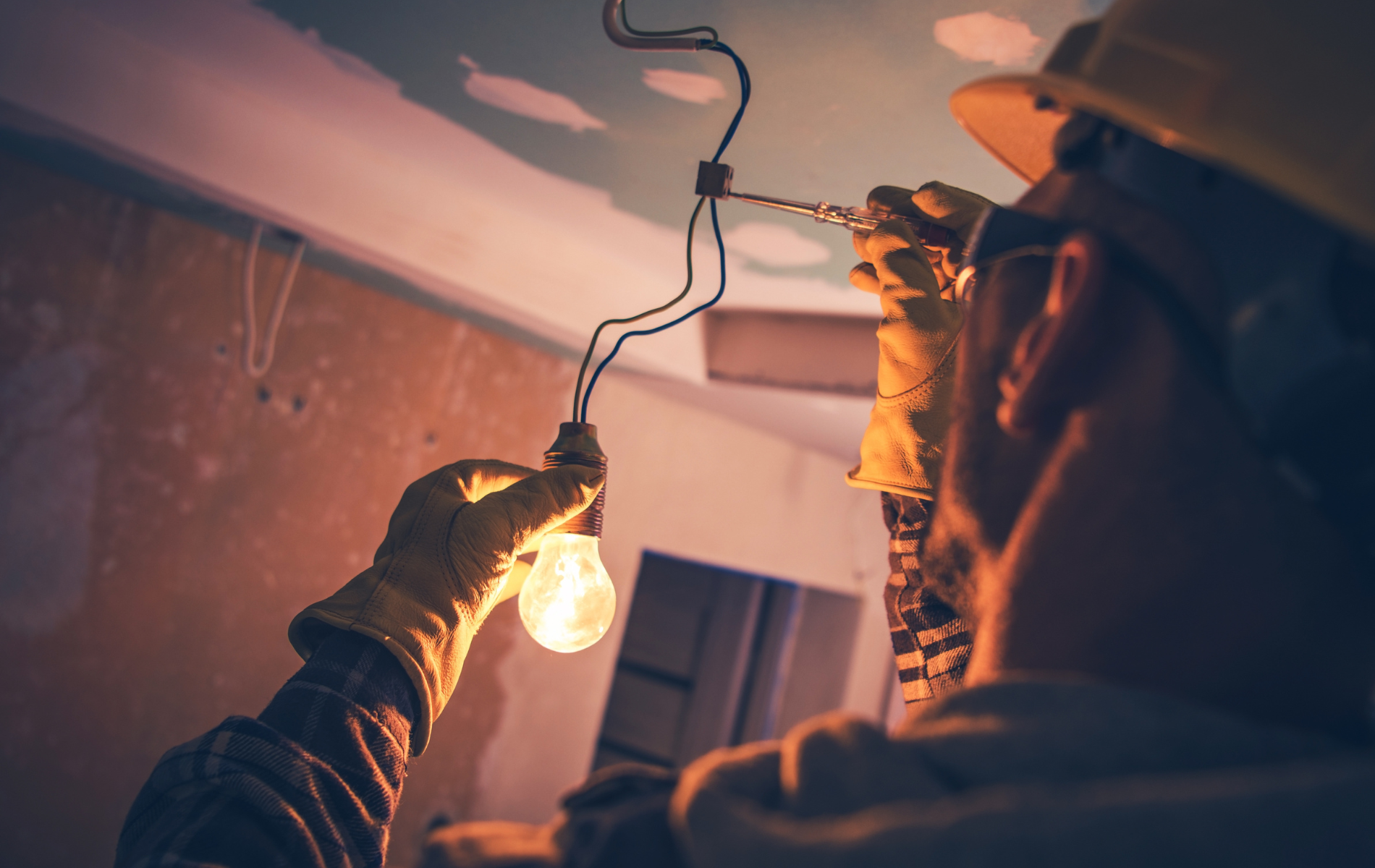 Electrician installing a lightbulb in a room with exposed wires, wearing gloves and a hard hat.