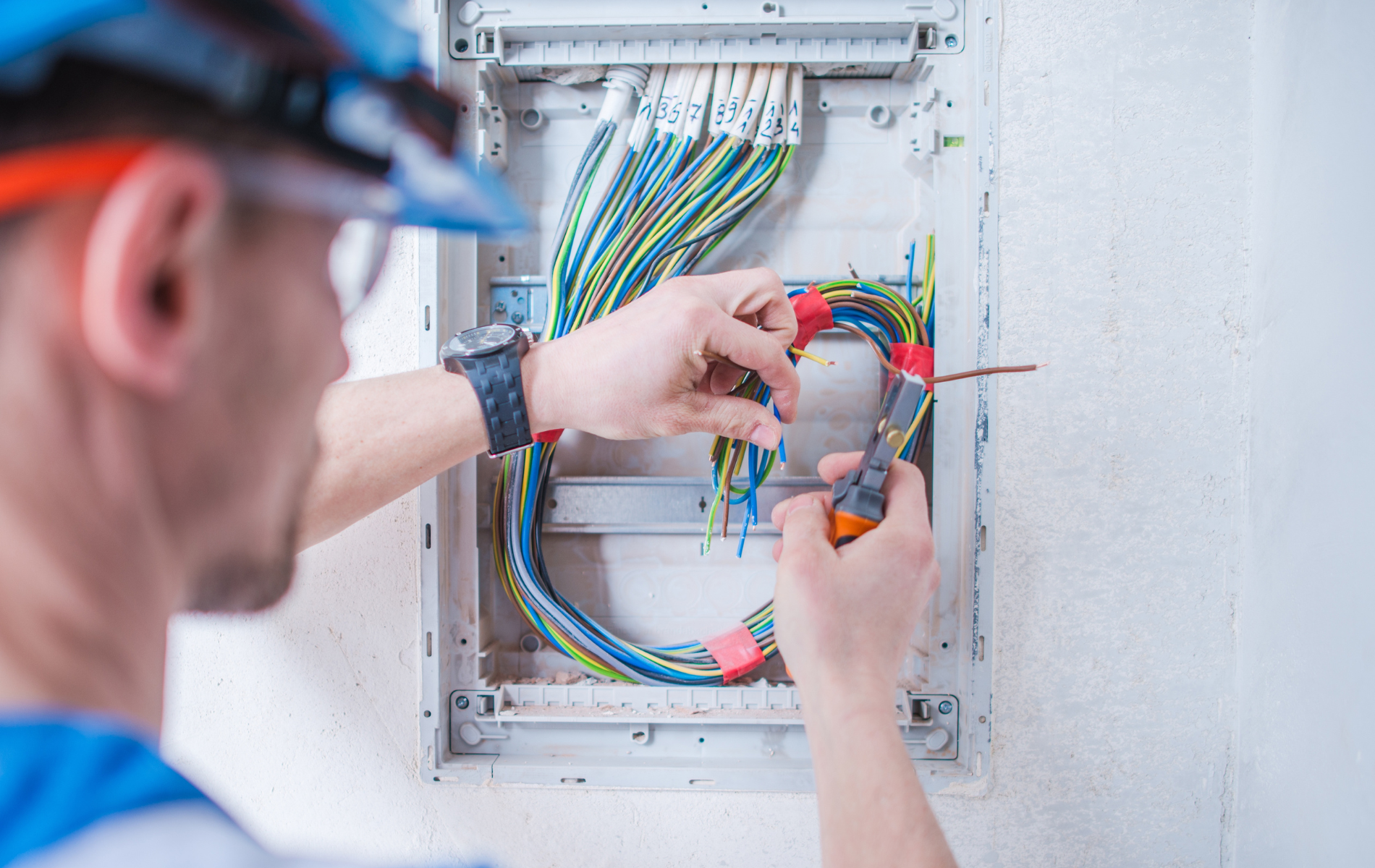 Electrician working on wiring inside a white electrical panel, wearing safety glasses and a blue hard hat.