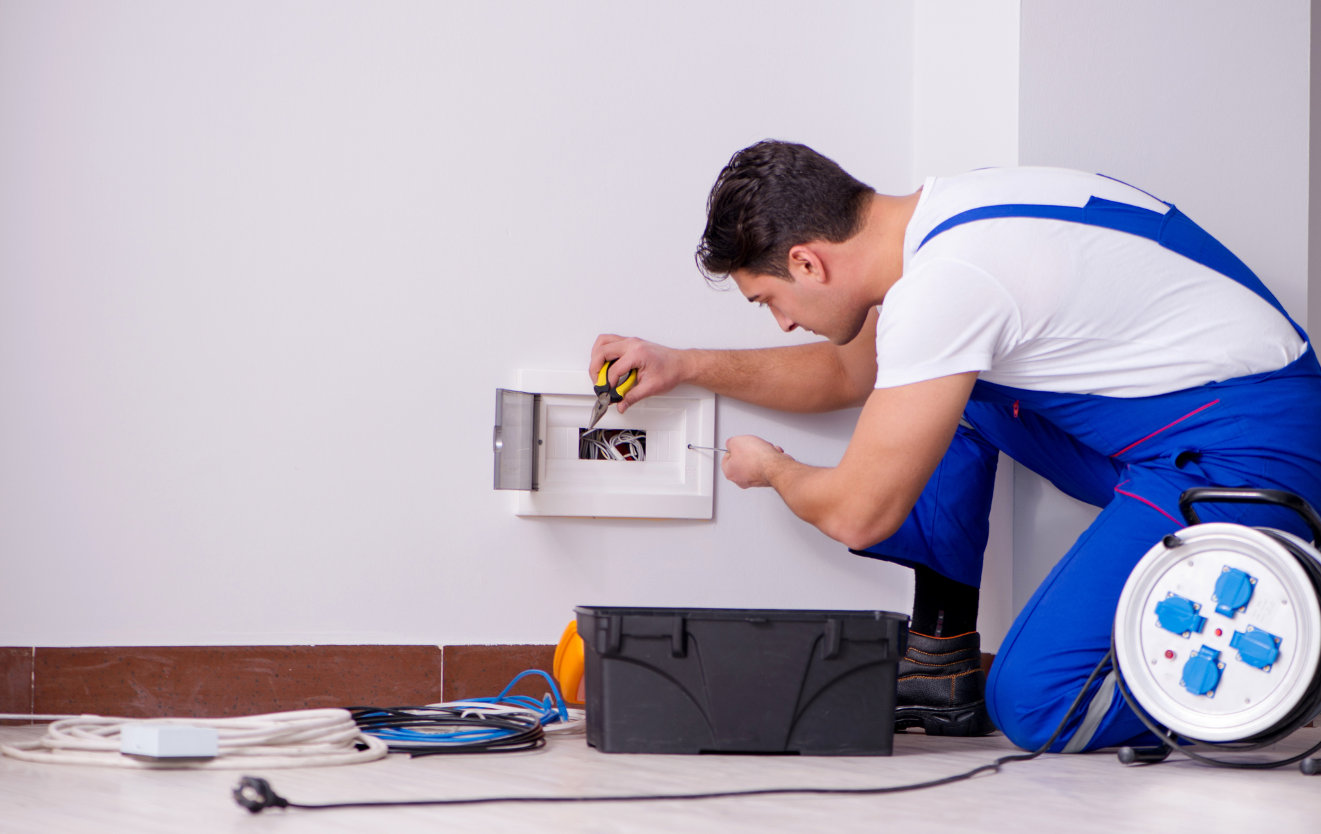 Electrician in blue coveralls repairs a breaker box on a white wall, with tools and an extension cord nearby.