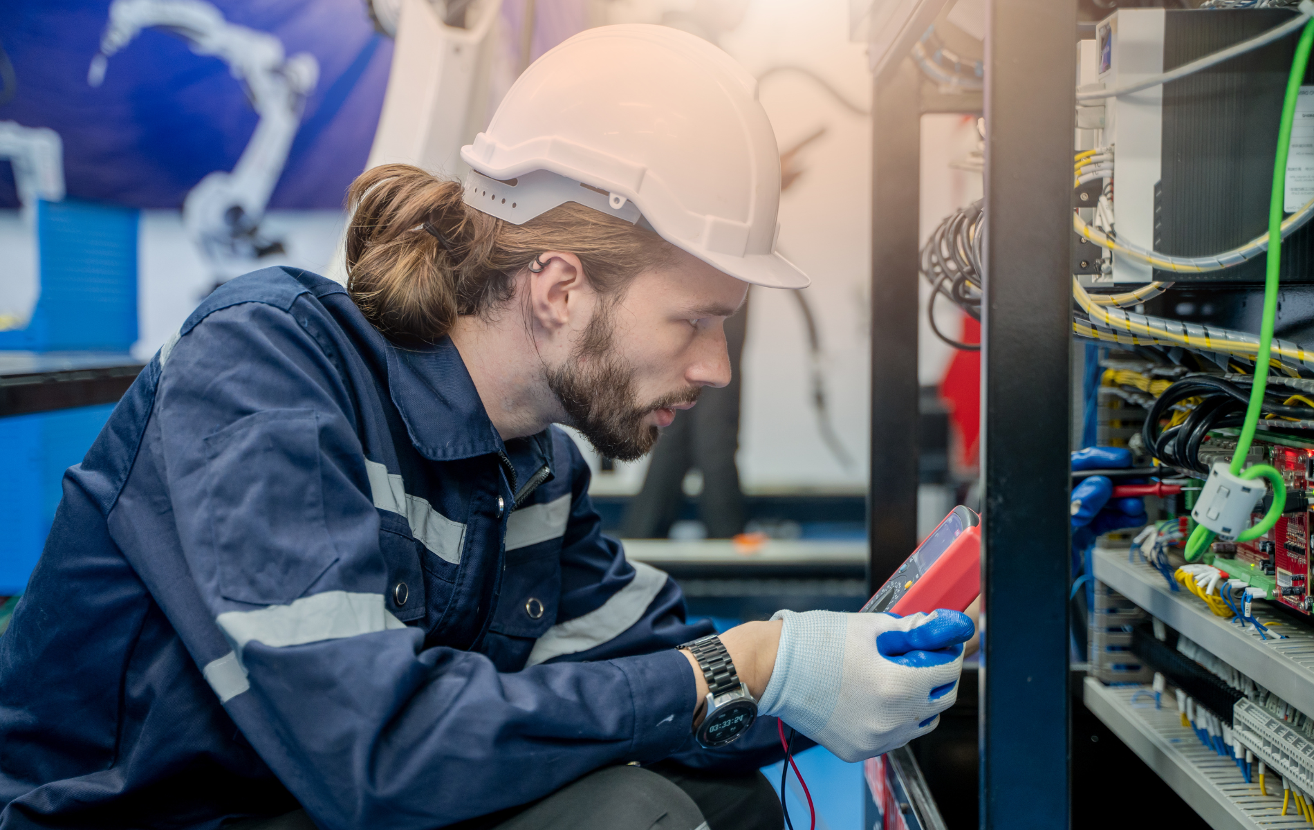Man in hard hat and gloves inspecting electrical panel with multimeter.