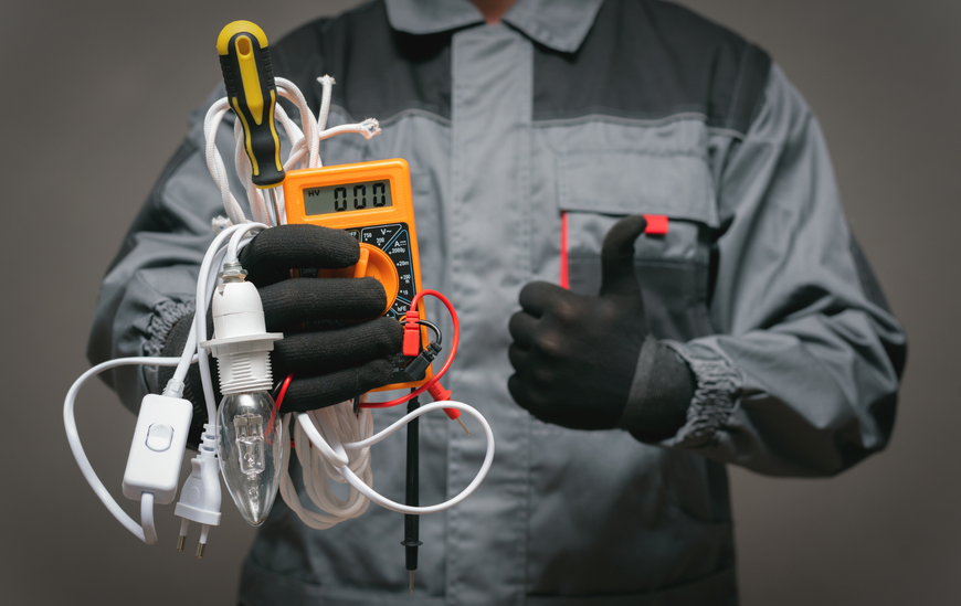 Electrician holding tools, wearing gloves, and giving a thumbs-up; grey background.