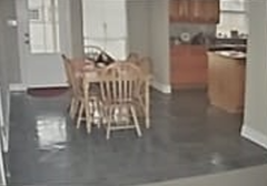 Grey-floored dining area with wooden table and chairs, leading to a kitchen with light-colored cabinetry.