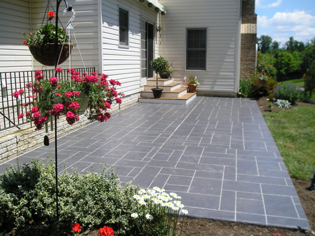 Grey stamped concrete patio next to a white house with flowers in hanging baskets and a garden.