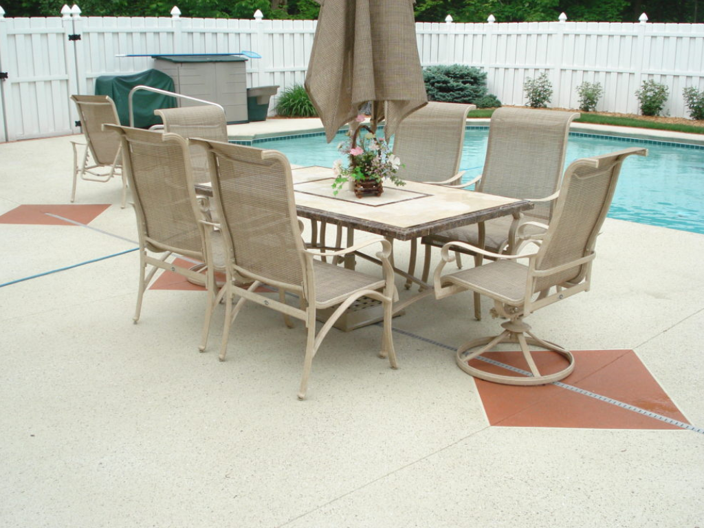 Patio table and chairs by a pool, with umbrella, white fence, and decorative squares.