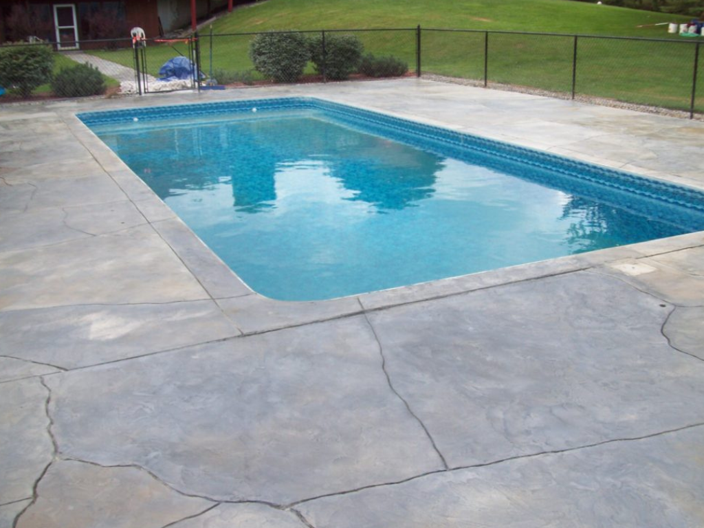 Rectangular in-ground swimming pool with blue water, surrounded by gray concrete patio and black fence on green grass.