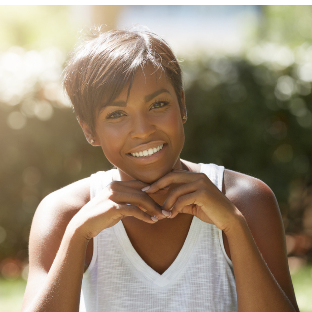 A woman in a white tank top smiles with her hands on her chin