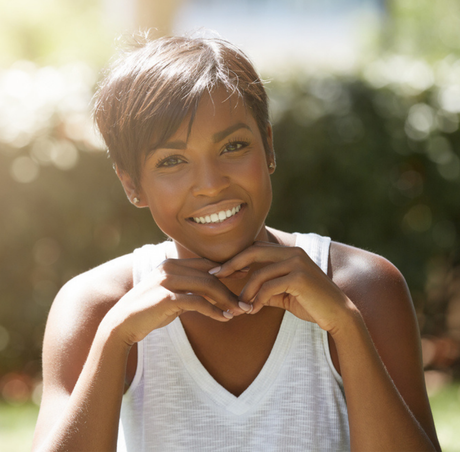 Smiling Black woman with short dark hair, outdoors, hands clasped near chin, wearing white tank top.