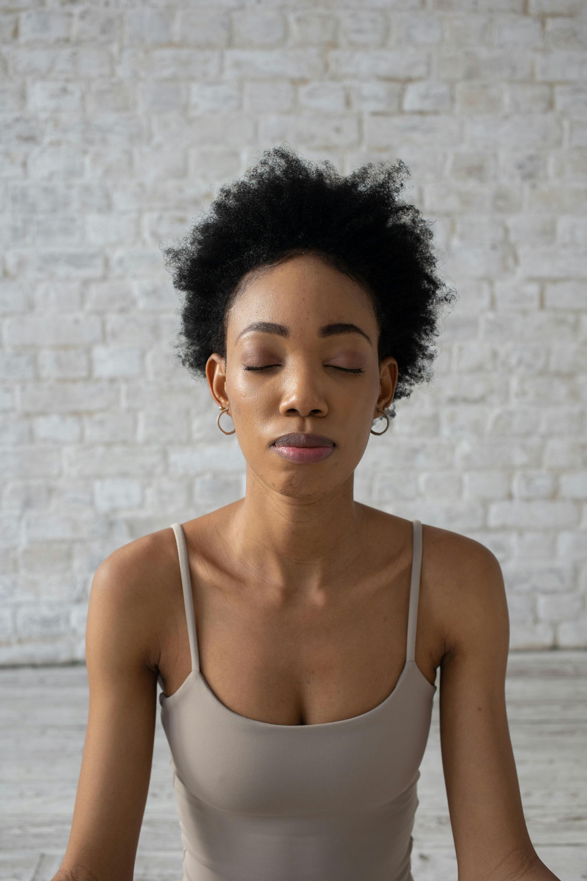 Woman in beige top meditating with eyes closed in front of a white brick wall.