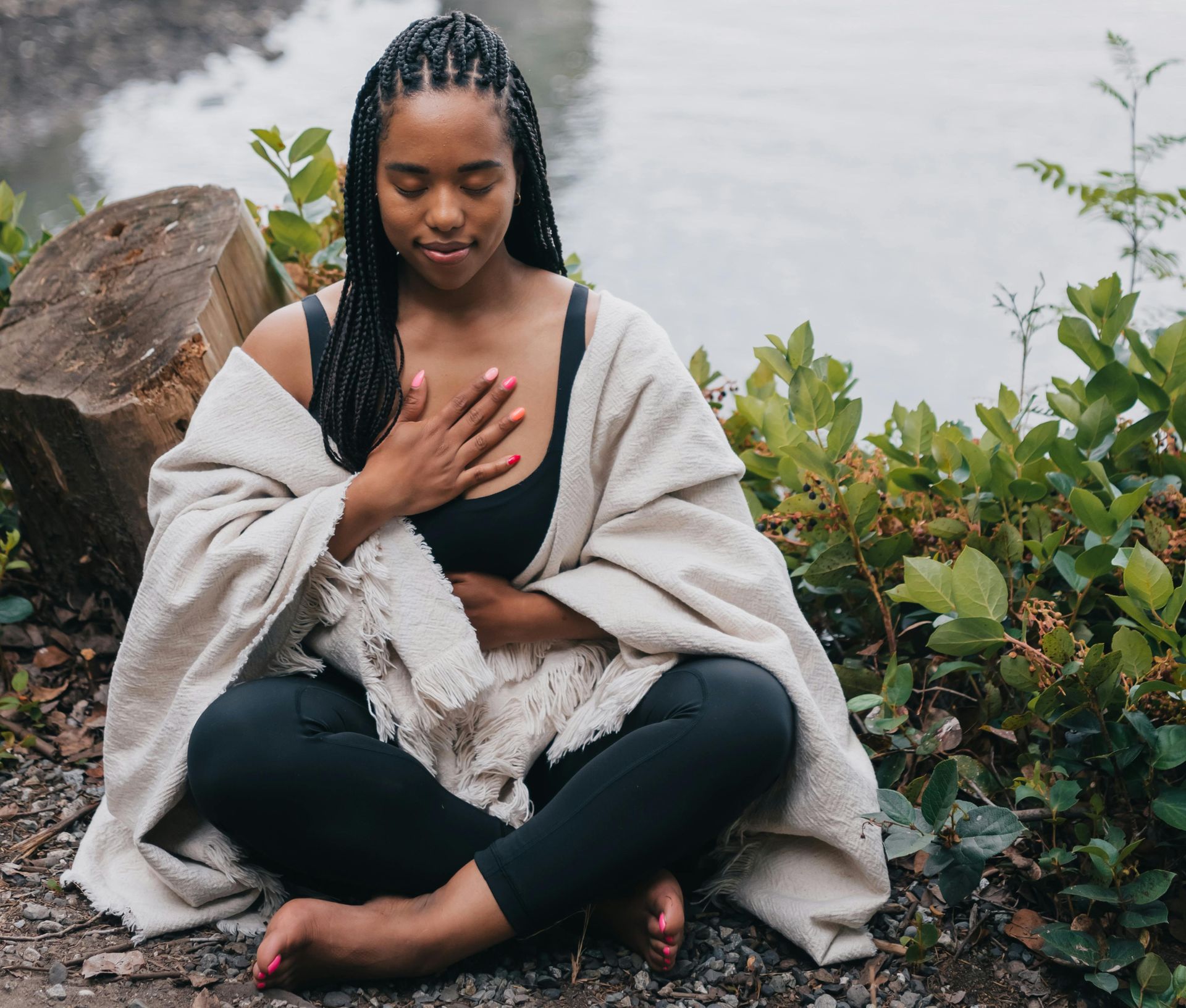 Woman in meditation pose, eyes closed, hand on chest, sitting near water, wrapped in a blanket.