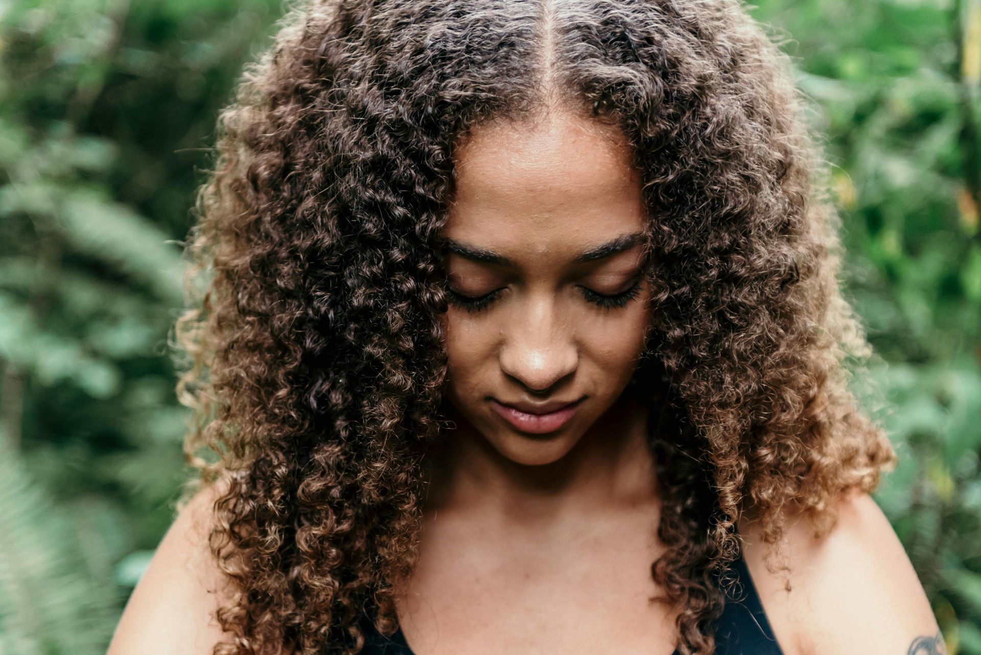 Woman with curly hair looking down outdoors with green foliage.